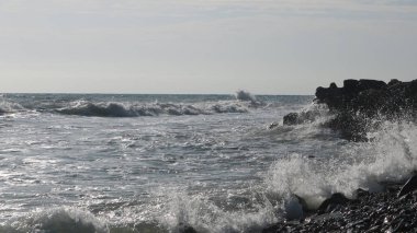 Gümüşi dalgalar ve köpüklü köpüklü deniz dalgaları güneşte ıssız sahildeki siyah kayalara vuruyor.