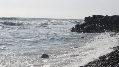 Suyun gümüş yansımasında köpüklü dalgalarla körfezin taştan kıyılarına vuran deniz sörfü. Işıltılı deniz dalgalarının arasından görünen ıssız kayalık kıyı şeridi.