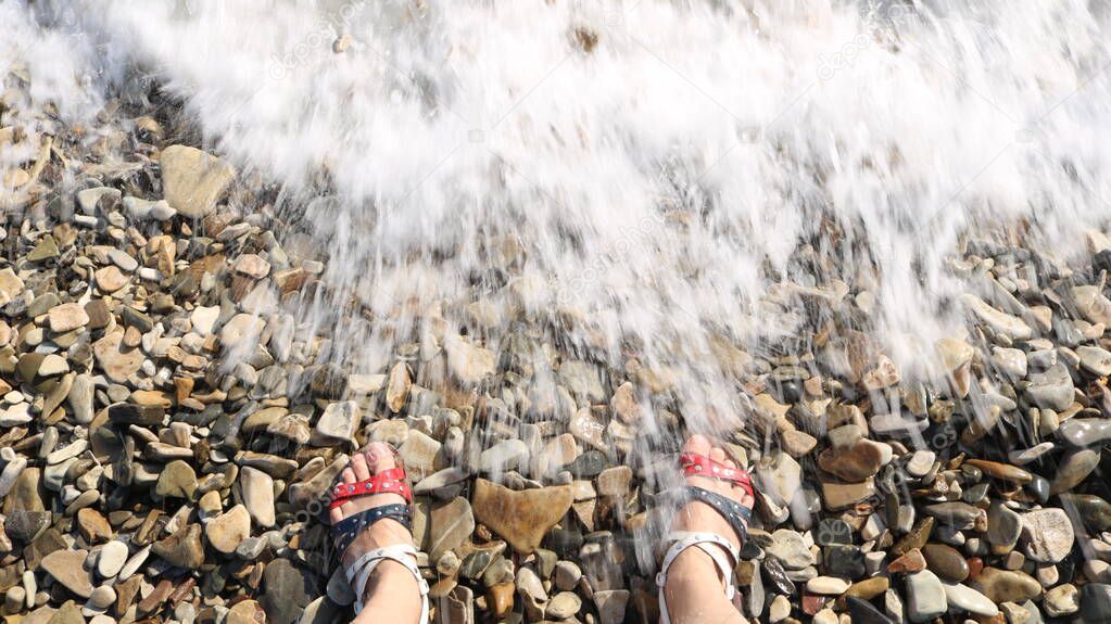 legs of a person in wicker sandals in American flag colors standing on a rocky beach in a spray of sea waves