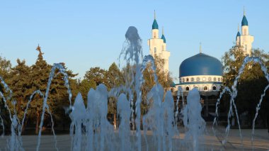 Maykop, Adygea / Rusya Cumhuriyeti - 5 Ekim 2020: Güneşli bir günde, mavi ve beyaz katedral camiinin arka planına karşı şehir meydanındaki bir fıskiyenin akışı ve sıçramaları
