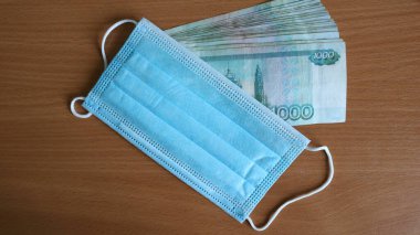 Bir Rus bankasının bininci banknotları mavi koruyucu bir tıbbi maskeyle birleşmiş, boş kahverengi bir masanın yüzeyinde duruyor, COVID 19 salgınına para akışının sembolü olarak.