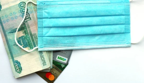 Rus parası bin banknot ve banka kartları beyaz bir arka planda, yukarıdan mavi bir tıbbi maske ile kaplanmış, bir salgın sırasında parasal ilişkilerin bir göstergesi.
