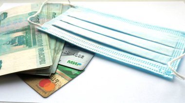 Bin ruble değerinde bir deste banknot, Visa ve Mir ödeme sistemlerinin kartları ve beyaz arka planda duran koruyucu bir tıbbi maske, bulaşıcı bir salgın sırasında para dolaşımını sembolize eden bir resim.