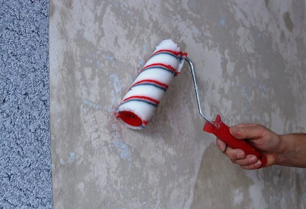 applying wallpaper glue with a striped roller on the wall in the process of gluing gray non-woven wallpaper, a man\'s hand with a red-white roller applying a layer of liquid glue to the prepared surface