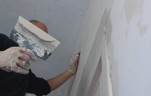 a male worker in dark clothes with a spatula at face level stands on a step-ladder against the wall and levels the slope after installing the door, a faceless plasterer with a placer in the process of repair work