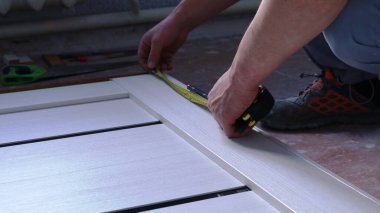 Bir marangozun elindeki mezurayı kullanarak yere metal bağlayıcılar yerleştirirken, yerde yatan hafif ahşap bir tuvalde ölçümler yapmak, bir odanın zemininde bir kapı çerçevesini birleştirmek,