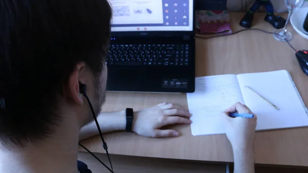 video class and online lectures for students via the Internet, a student making notes in a notebook at his home table while sitting in front of a laptop screen with headphones, teaching using technology