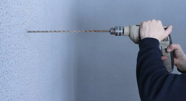 a hammer drill with a long attachment in the hands of a black-clad master, ready to drill a hole in a concrete wall covered with gray textured wallpaper, an installer with a tool in profile