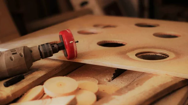 an electric drill with a red nozzle with a crown lying on a large wooden board in which round holes were cut, a pause in the carpentry workshop with a set aside tool and an exclusive product in the manufacturing process