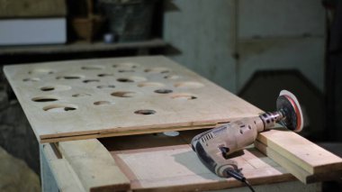 Bir marangozluk atölyesinde duraklatılmış yuvarlak deliklerle birlikte, usta tarafından ahşap bir tuval üzerine yerleştirilmiş zümrüt bir eklentiye sahip bir matkap, masaüstünde elle tutulan bir elektrikli alet