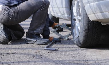 Bir adam evdeki gümüş bir arabanın tekerleğini asfalt bir yolda tekerlek değiştiren küçük bir kriko ve el aletinin yardımıyla değiştirecek.