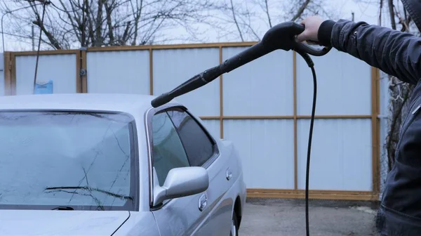 high-pressure washer in the process of cleaning a gray car in a private yard, a man's hand with a gun with a pressure of water washes the silver body of the car, home car wash