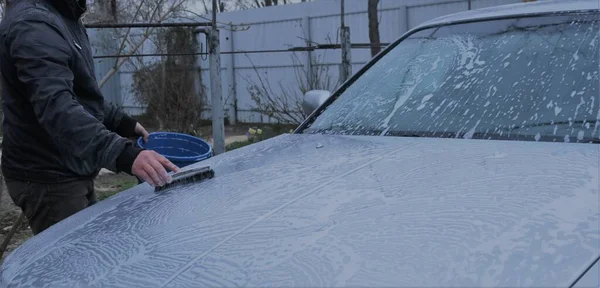 hand washing the hood of the car with a brush with soap and water from a bucket, home care of the car, soap foam on the surface of the car in the process of cleaning by a man at home