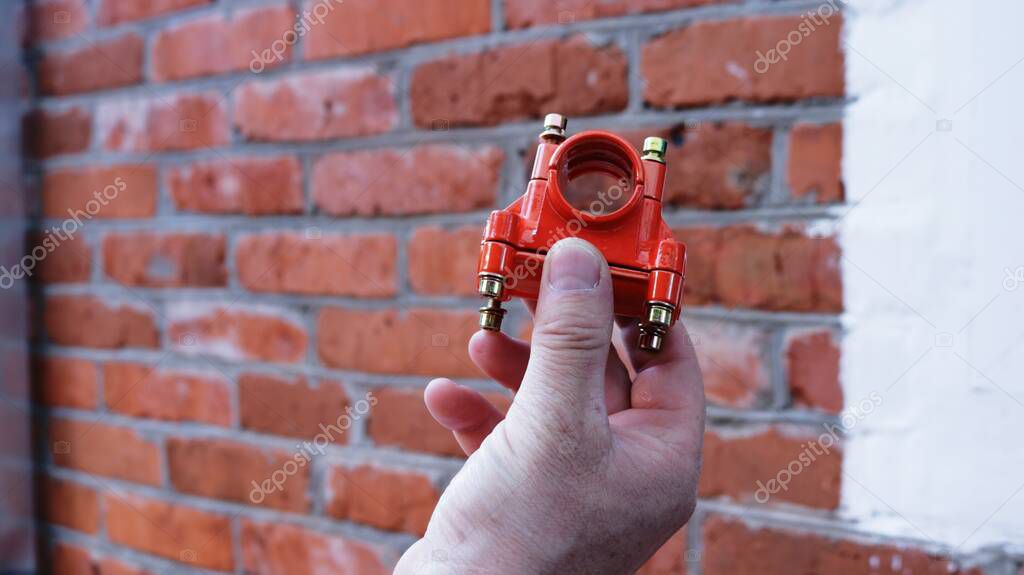 a man's hand holding an orange metal crane from a lawn mower close-up, demonstrating a useful part from a mower against a brick wall background, part of a fastener in the structure of a gasoline mower