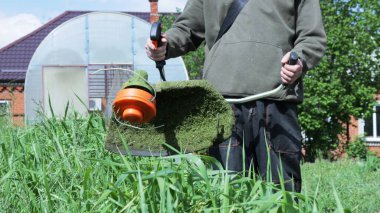 Bahçedeki binaların arka planına karşı kırsal bir alanda çalışan bir aletle çim biçme makinesi, yaz mevsiminde çimleri elle biçmek için bir benzin biçme makinesi kullanılıyor.
