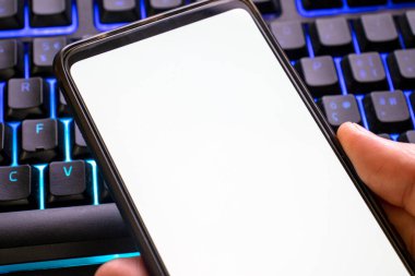 Elinde beyaz beyaz ekranla cep telefonunu taklit et. arkaplanda mavi ışıklı klavye ile boş ekran ile akıllı telefon modellemesi