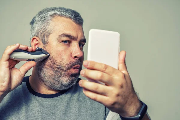 gray-haired man cuts his beard with an electric clipper