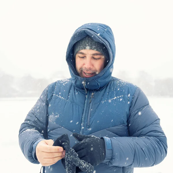 A 40-year-old man in a blue down jacket and a hood puts on gloves. blizzard and cold. face is not visible due to snow