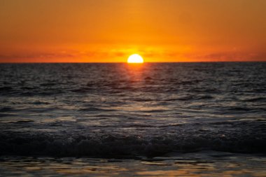 Güneş, koyu kırmızı bir günbatımında denize batıyor. Karanlık bir denizde dalgaların hareketini görebiliyoruz.