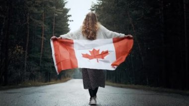 Beyaz kazak ve etekli bir kız elinde Kanada bayrağıyla ormanda yürüyor.