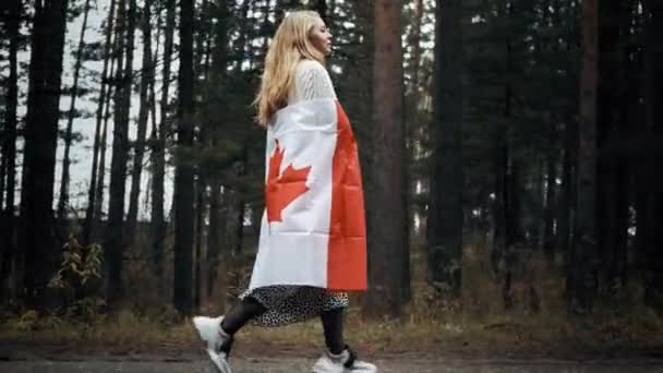 Een meisje in een witte trui en rok loopt door het bos met de vlag van Canada, zijaanzicht — Stockvideo