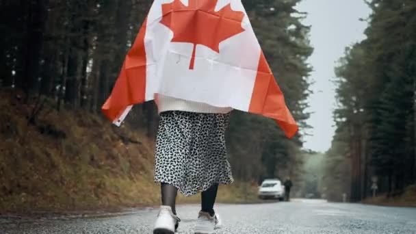 Een meisje in een witte trui en rok loopt door het bos met de vlag van Canada — Stockvideo