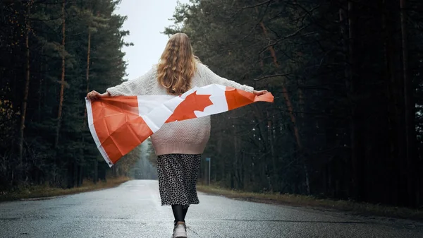 Ένα κορίτσι με λευκό πουλόβερ και φούστα περπατά μέσα στο δάσος κρατώντας τη σημαία του Καναδά — Φωτογραφία Αρχείου