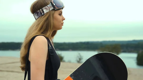 Chica en overoles y gafas en los paseos de verano en la arena con una tabla de snowboard —  Fotos de Stock