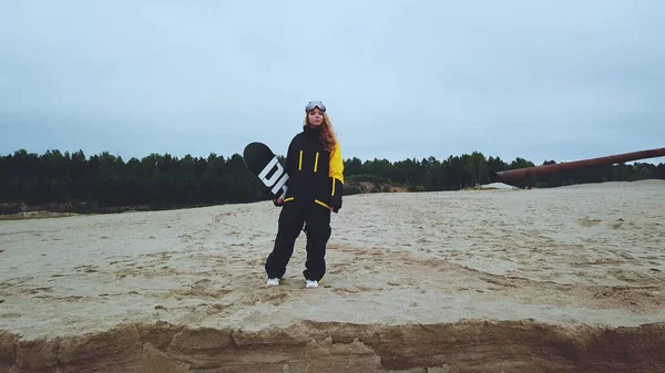 Una chica en overol y gafas en el verano mira al lago sosteniendo una tabla de snowboard en sus manos —  Fotos de Stock