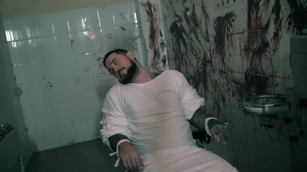 Homem louco de camisa de força amarrado a uma cadeira, psicopata assassino em uma sala com overdose de paredes ensanguentadas — Fotografia de Stock