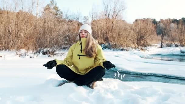 La niña se sienta en la posición de loto en el invierno en el bosque y mira al río. Vista frontal — Vídeos de Stock