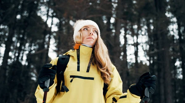 Menina com um chapéu branco na floresta com uma mochila e caminhadas varas no inverno. Vista frontal. Fechar — Fotografia de Stock