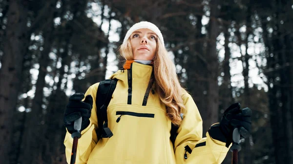 Chica con un sombrero blanco en el bosque con una mochila y bastones de senderismo en invierno. Vista frontal. De cerca. —  Fotos de Stock