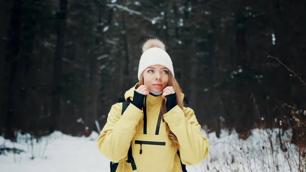 Menina com um chapéu branco na floresta com uma mochila no inverno endireita seu casaco. Vista frontal — Fotografia de Stock