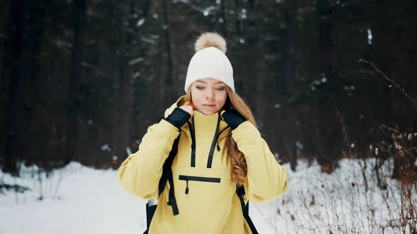 Menina com um chapéu branco na floresta com uma mochila no inverno endireita seu casaco. Vista frontal — Fotografia de Stock