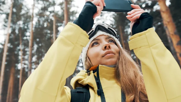 Дівчина в білому капелюсі і сноуборд маска в лісі фотографує природу на телефоні взимку — стокове фото