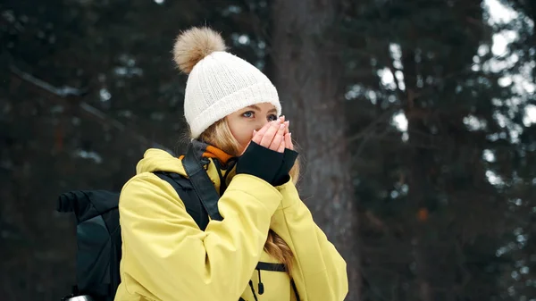 Uma menina de chapéu branco e um anorak amarelo aquece as mãos no inverno na floresta — Fotografia de Stock