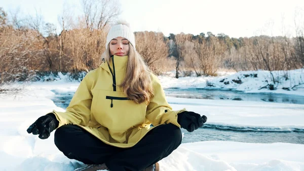 A menina se senta na posição de lótus no inverno na floresta e olha para o rio. Vista frontal — Fotografia de Stock