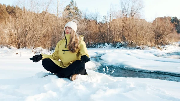 A menina se senta na posição de lótus no inverno na floresta e olha para o rio. Vista frontal — Fotografia de Stock