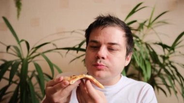 Genç adam bir dilim pizza alıyor, tutkuyla ısırıyor ve çiğniyor. Adam bir dilim pizzaya hayranlıkla bakıyor. Lezzetli peynirli pizza. Arka planda yeşil bitkiler.