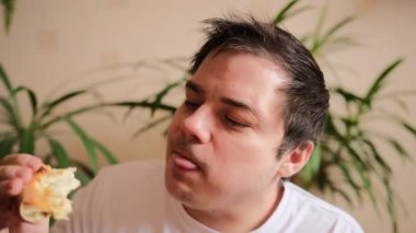 Beyaz tişörtlü karizmatik bir adam lezzetli peynirli pizza yer ve büyük bir zevkle gazozla içer. Genç adam bir dilim pizza alıyor, bir ısırık alıyor ve çiğniyor. Yakın plan. Arka planda yeşil bitkiler.