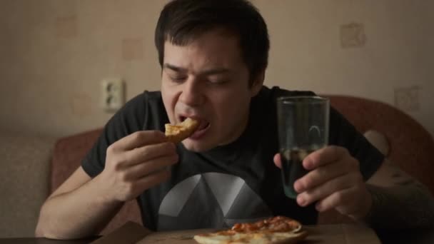 黒いTシャツの若い男がおいしいピザを食べ 彼の手にソーダとガラスを保持しています 男はピザのスライスを保持します 一口食べて それをかむ — ストック動画