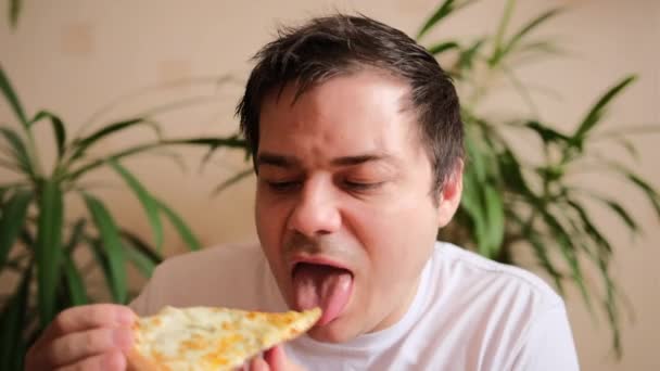 白いTシャツの素敵な男は大きな喜びとおいしいチーズピザを食べます 若い男は ピザのスライスを取ります 一口を取り それを噛む 接近中だ 背景にある緑の植物 — ストック動画
