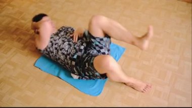 Şortlu ve tişörtlü bir adam yerde yatarken karın kaslarını sallıyor. Bir adam karın kasları üzerinde egzersiz yapıyor. Evde spor aktiviteleri. Şişman adam kilo vermek için aktif olarak sporla ilgileniyor. Ev çalışması.