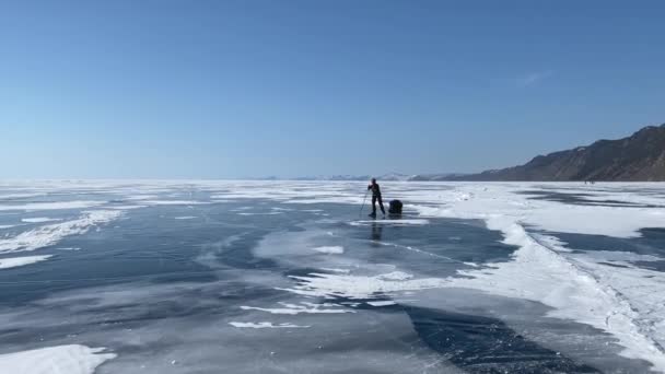 一名戴头盔的女运动员 溜冰鞋 膝盖垫 滑雪杆和拖着背包在冰冻的贝加尔湖冰上骑车 美丽的冬季风景 蓝色透明的冰 有深深的裂缝和雪岛 — 图库视频影像