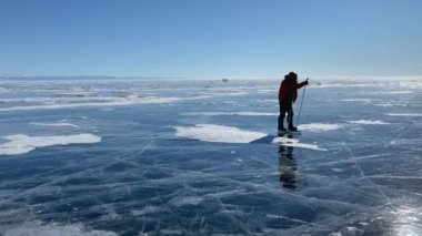 Kırmızı kabarık ceketli ve turuncu kasklı bir adam donmuş Baykal Gölü 'nün buzunda kayıyor. Güzel kış manzarası berrak, pürüzsüz mavi şeffaf buz derin çatlaklar ve kar parçalarıyla.