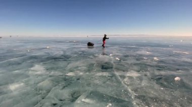 BAIKAL, IRKUTSK REGION, RUSSIA - 18 Mart 2021: Bir kadın donmuş Baykal Gölü 'nün buzunda paten üzerinde seyahat eder. Arkadan bak. Derin çatlakları olan saydam mavi buzla güzel bir kış manzarası..
