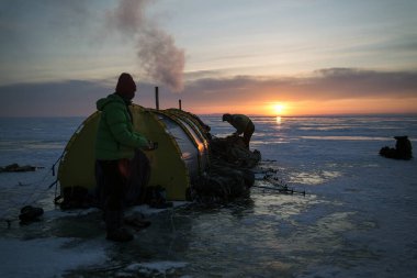 Turistler, donmuş karla kaplı Baykal Gölü kıyısında bir sobayla bir kış çadırında duruyorlar. Güzel gün batımı. Kış yürüyüşü. Kuzey manzarası. Rusya 'nın doğal simgesi.