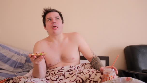 裸の胴を持つけだもの太った男がベッドに座り テレビを見てピザを食べる 若い男はピザを一口食べて噛み ミルクセーキを手にして — ストック動画