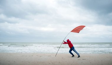 On the shore of stormy sea person struggling with the wind. The red flag indicates the strength of the wind. clipart